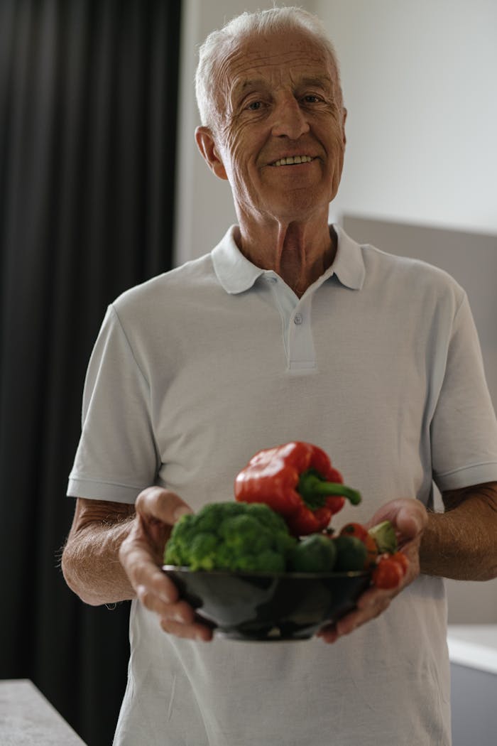 services-04 Elderly man happily holding a bowl of fresh vegetables indoors, promoting healthy lifestyle.